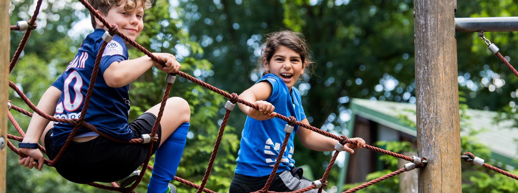 Zwei Kinder spielen auf einem Klettergerüst aus Seilen in einem Park, sie lachen und haben Spaß.