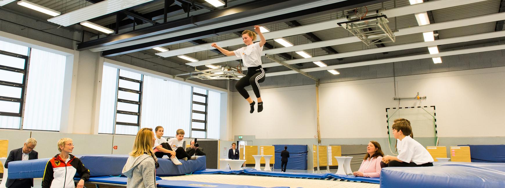 Junge springt auf einem Trampolin, während Zuschauer und Trainer im Hintergrund beobachten. Sporthalle mit hellem Licht.